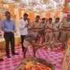 Group of men, including police officers, standing around a ritual fire and offerings at a decorative ceremony outdoors with orange decorations and garlands.