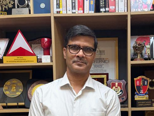 Man with glasses in a light striped shirt stands in front of shelves filled with trophies, plaques, and books.