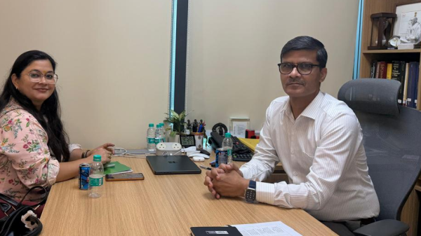 Two professionals sit at a wood desk in an office, facing the camera; laptop, water bottles, and office supplies are on the desk.