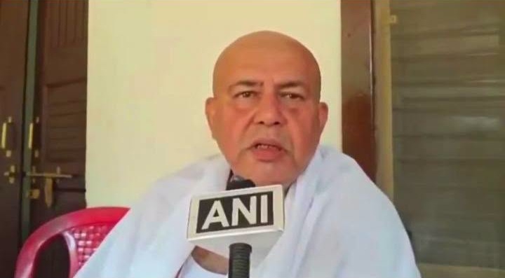 Bald man wearing white clothing speaks into an ANI microphone during an interview, seated indoors with a red chair behind him.