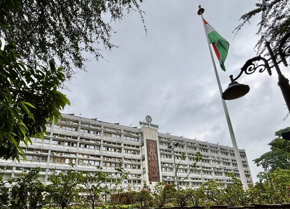 Front view of a large white multi-story building with a tall flagpole flying the Indian flag, surrounded by trees and a decorative street lamp against a cloudy sky.