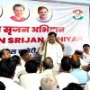 Man in a white kurta addressing a seated audience at a political rally, banner with portraits and a hand party symbol behind him.