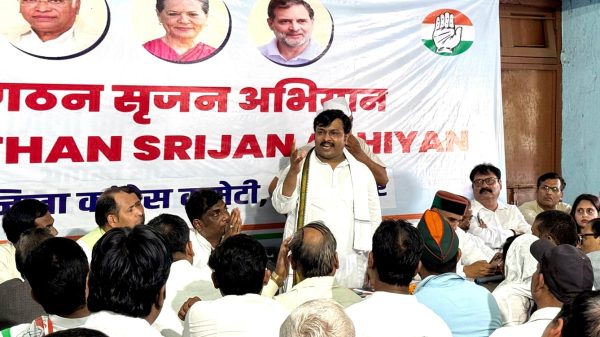 Man in a white kurta addressing a seated audience at a political rally, banner with portraits and a hand party symbol behind him.