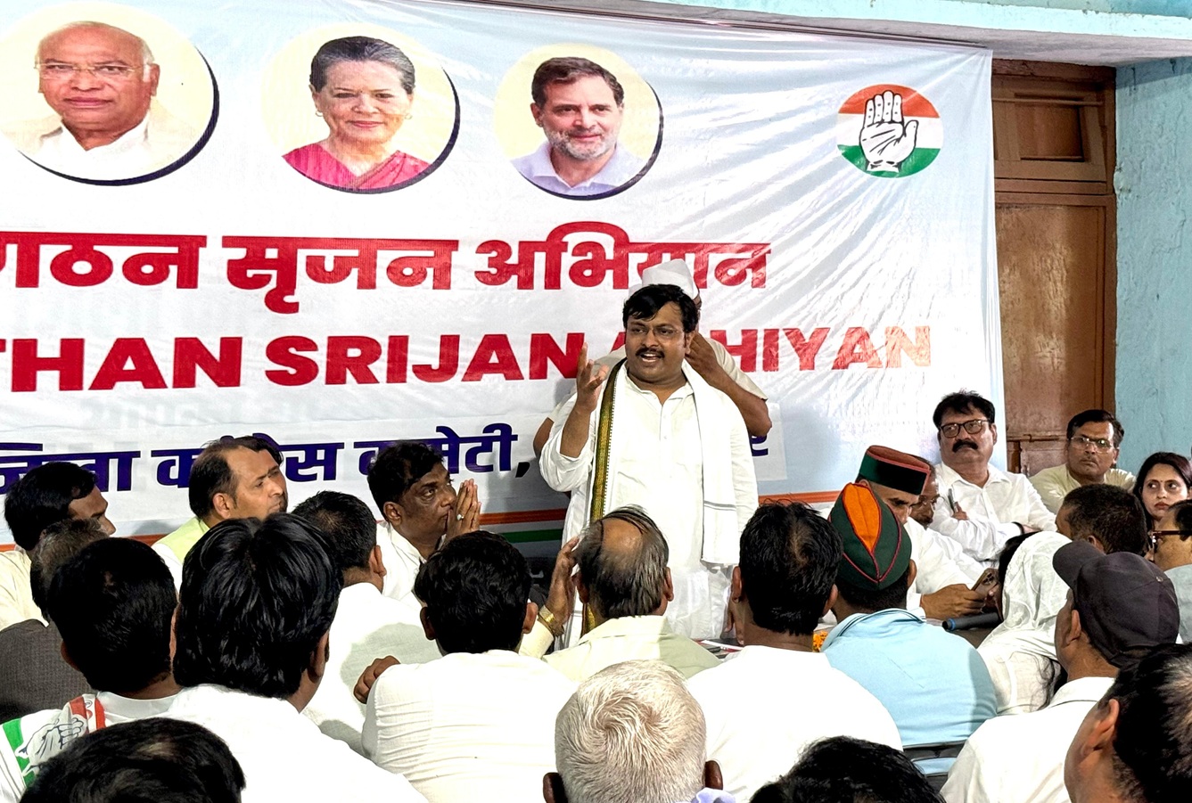 Man in a white kurta addressing a seated audience at a political rally, banner with portraits and a hand party symbol behind him.