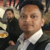 Selfie of a man in a white shirt and dark vest at a social gathering, with guests mingling and a person eating in the background.