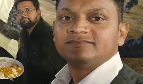 Selfie of a man in a white shirt and dark vest at a social gathering, with guests mingling and a person eating in the background.
