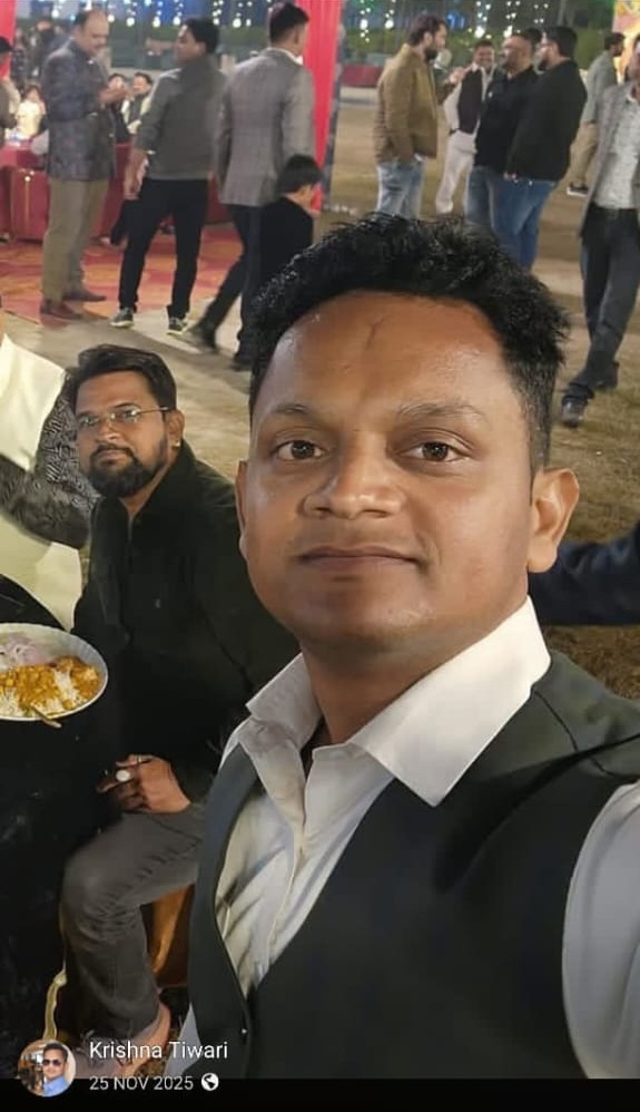 Selfie of a man in a white shirt and dark vest at a social gathering, with guests mingling and a person eating in the background.