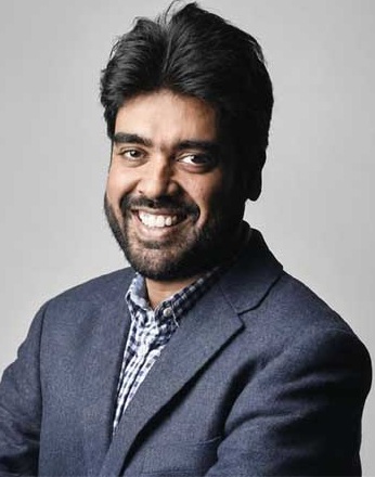 kunal pradhan | No. 1 Indian Media News Portal Smiling man with dark hair and beard wearing a navy blazer over a checkered shirt on a gray backdrop.
