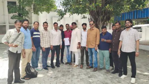 Group of men standing in a line outside on a paved courtyard under trees, posing for a photo.