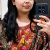 Woman taking a mirror selfie with a dark phone, wearing a colorful embroidered traditional outfit and sunglasses on her head.
