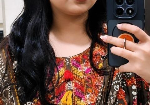 Woman taking a mirror selfie with a dark phone, wearing a colorful embroidered traditional outfit and sunglasses on her head.
