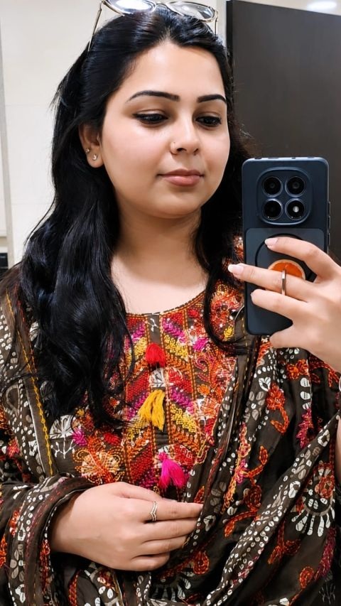 Woman taking a mirror selfie with a dark phone, wearing a colorful embroidered traditional outfit and sunglasses on her head.