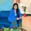 Woman in a blue suit sits on a blue sofa in a lobby, with potted plants on a glass table nearby.