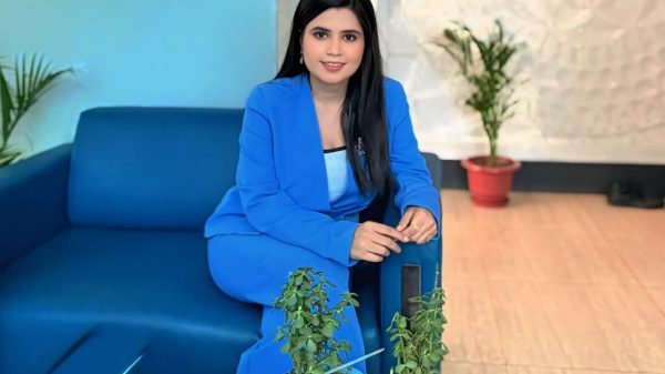 Woman in a blue suit sits on a blue sofa in a lobby, with potted plants on a glass table nearby.