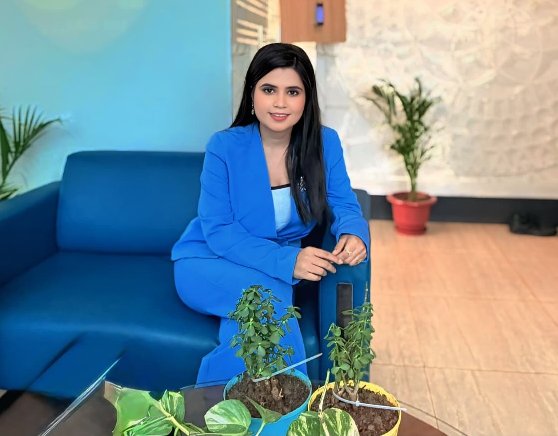 Woman in a blue suit sits on a blue sofa in a lobby, with potted plants on a glass table nearby.
