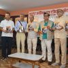 Seven men in casual and semi-formal attire pose with booklets at a book launch, banner in Hindi behind them.