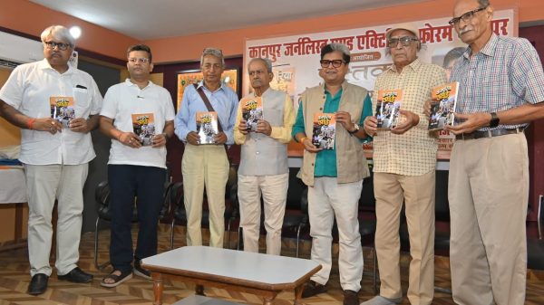 Seven men in casual and semi-formal attire pose with booklets at a book launch, banner in Hindi behind them.