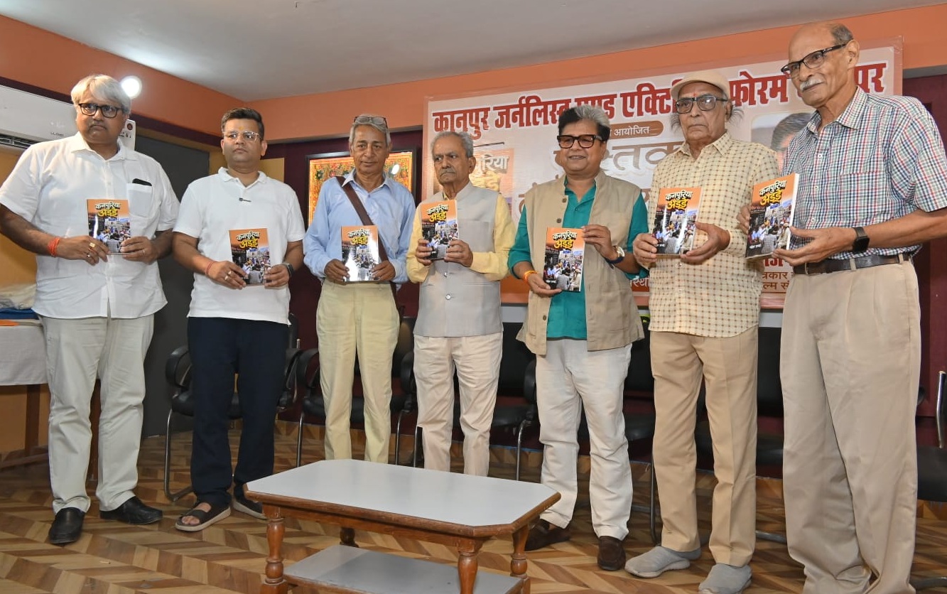 Seven men in casual and semi-formal attire pose with booklets at a book launch, banner in Hindi behind them.