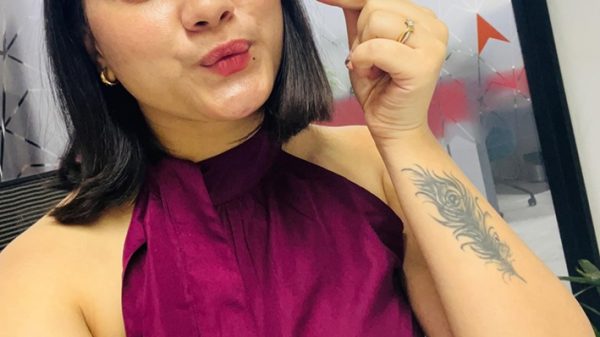 Woman with shoulder-length black hair posing for a selfie in an office, wearing a burgundy sleeveless blouse and a ring on her finger, with a forearm tattoo visible on her right arm.