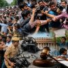 Collage of protests with crowds clashing with police, journalists filming, a student holding a blank sign, and symbols of government and judiciary in the background.