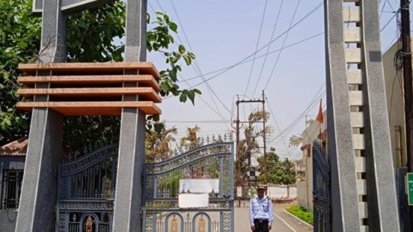 Entrance gate with tall grey pillars and a sign in Hindi reading 'Media City'; a security guard stands on the road inside the gate.