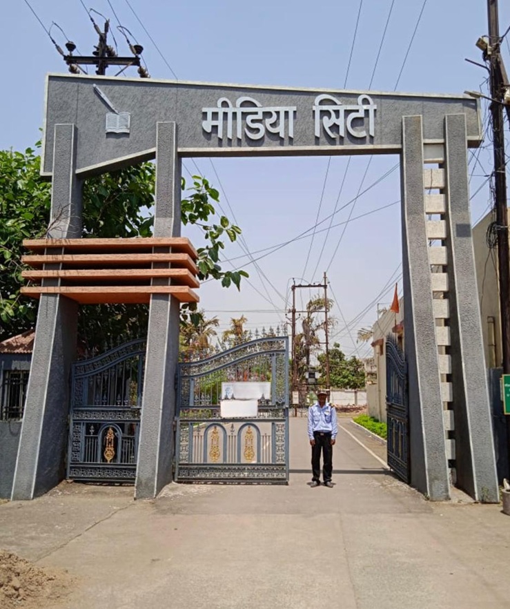 Entrance gate with tall grey pillars and a sign in Hindi reading 'Media City'; a security guard stands on the road inside the gate.