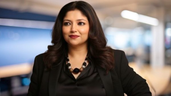 Professional woman with shoulder-length dark hair wearing a black blazer and beaded necklace in a modern office setting, looking to the side.
