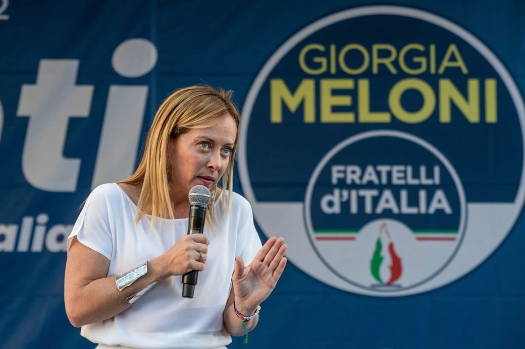 Woman in a white blouse speaks into a handheld microphone on a stage, with a large blue campaign banner and circular logo in the background.
