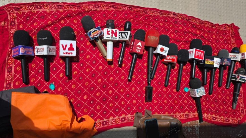 mike down_1 | No. 1 Indian Media News Portal Row of handheld microphones with various news logos arranged on a red patterned cloth, ready for a press briefing.