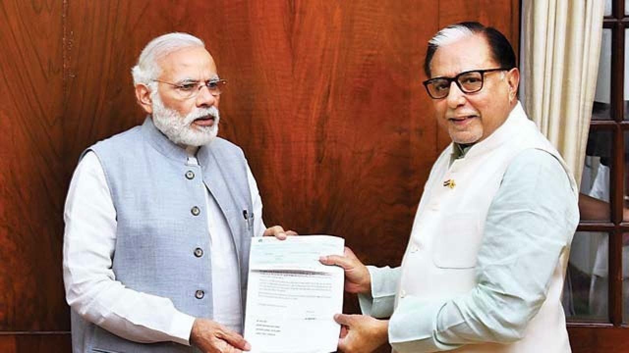 Two men exchanging a document indoors; one in a light gray vest and white kurta, the other in a white outfit, both smiling.