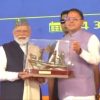 Elderly man with white beard and gray cap receives a glass trophy from a man in a lavender vest on a blue stage as others applaud.