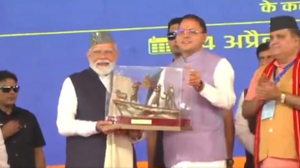 Elderly man with white beard and gray cap receives a glass trophy from a man in a lavender vest on a blue stage as others applaud.