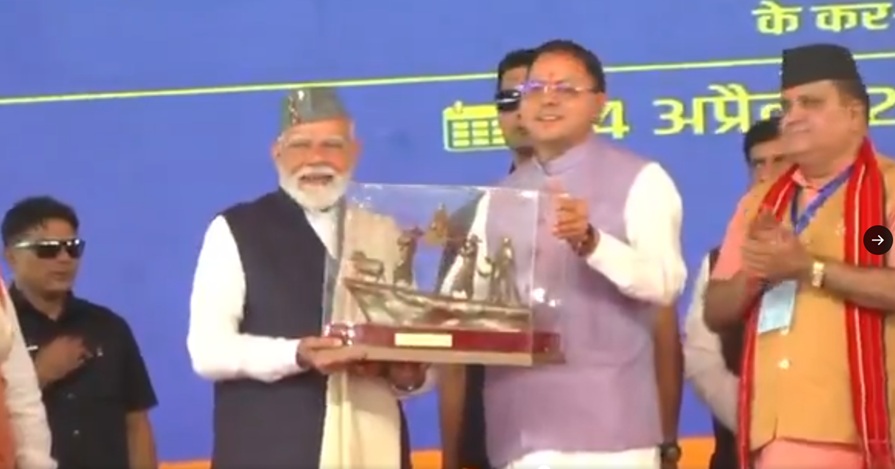 Elderly man with white beard and gray cap receives a glass trophy from a man in a lavender vest on a blue stage as others applaud.