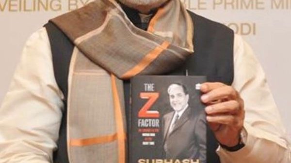 Older man with white hair and beard, wearing a scarf and vest, holds a book with a black cover showing a portrait and the title 'The Z Factor'.