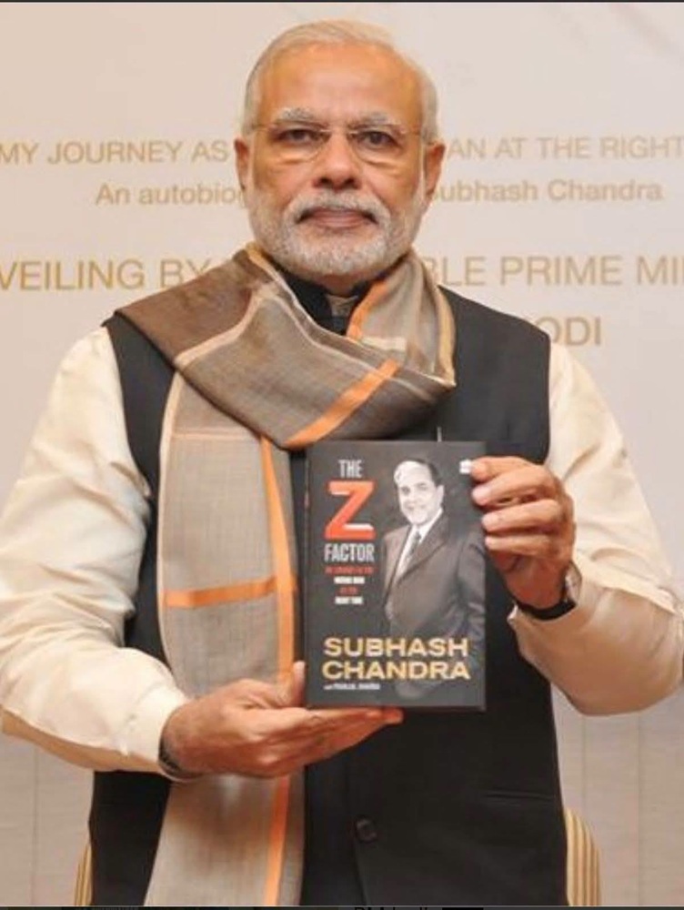 Older man with white hair and beard, wearing a scarf and vest, holds a book with a black cover showing a portrait and the title 'The Z Factor'.