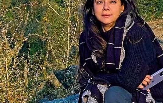 Woman with long dark hair sitting on a mossy rock outdoors, wearing a dark sweater and scarf, looking at the camera.