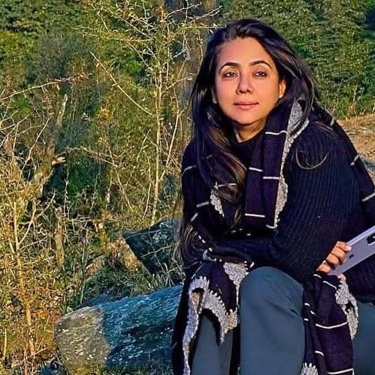 Woman with long dark hair sitting on a mossy rock outdoors, wearing a dark sweater and scarf, looking at the camera.