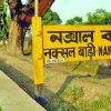 Yellow signboard with Bengali text reading 'Naksal Bari' beside a tree-lined sidewalk in a park area.