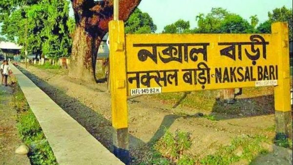 Yellow signboard with Bengali text reading 'Naksal Bari' beside a tree-lined sidewalk in a park area.