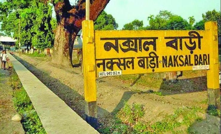 Yellow signboard with Bengali text reading 'Naksal Bari' beside a tree-lined sidewalk in a park area.
