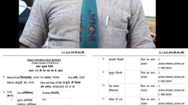 Man wearing glasses speaks into a blue microphone labeled 'INDIA' during a news report and papers in the background.