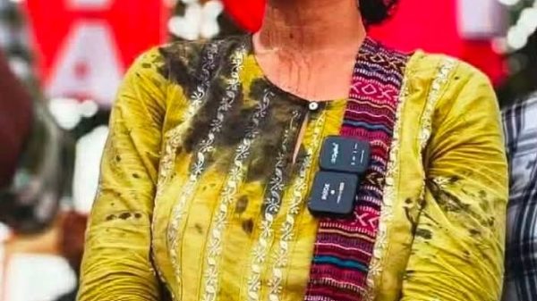 Woman in a yellow traditional kurta standing with folded arms at an outdoor event, wearing a woven strap with badges draped across her chest.