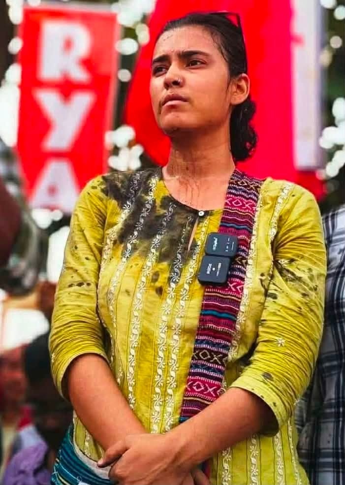 neha aisa | No. 1 Indian Media News Portal Woman wearing a yellow kurti with a patterned crossbody strap, standing with folded arms at an outdoor event with red banners in the background.
