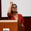 Woman in red blouse with glasses speaking at a podium, raising her index finger into the air beside a microphone.