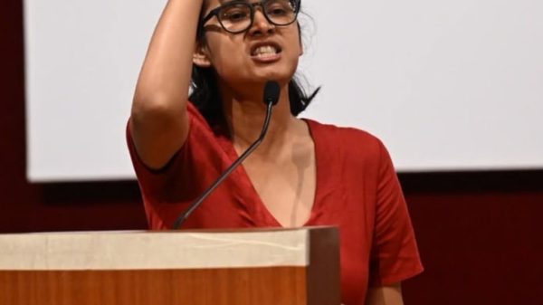 Woman in red blouse with glasses speaking at a podium, raising her index finger into the air beside a microphone.