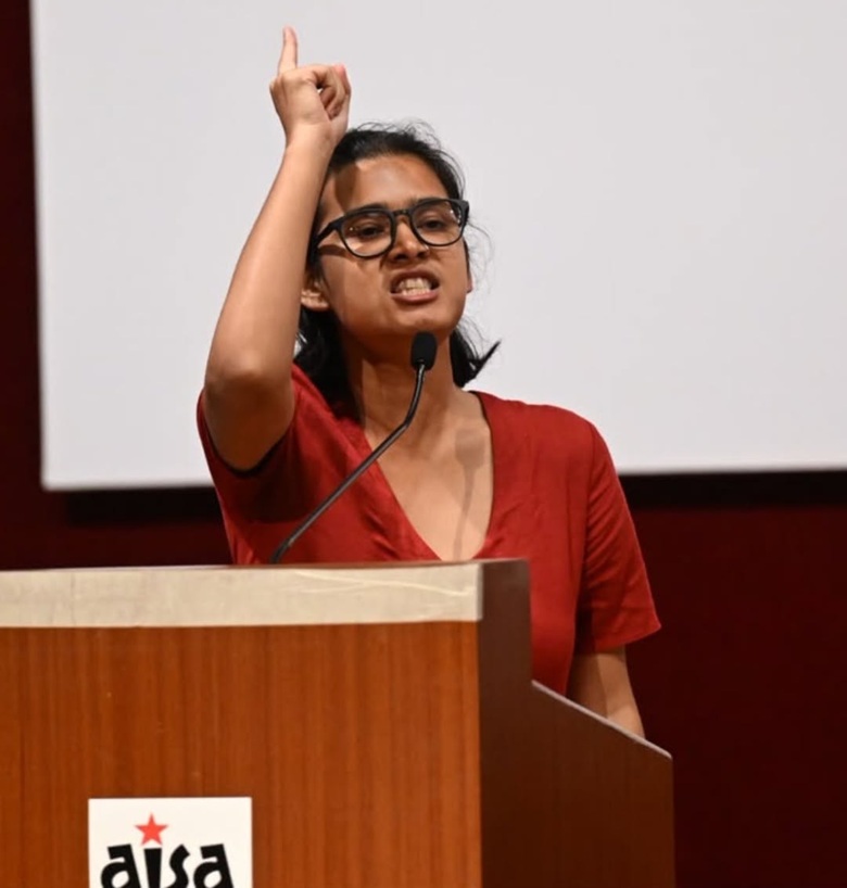 Woman in red blouse with glasses speaking at a podium, raising her index finger into the air beside a microphone.