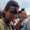 Young woman with glasses and a yellow shirt, mud splattered on her face and clothes, smiling in a crowd as someone takes a selfie nearby.