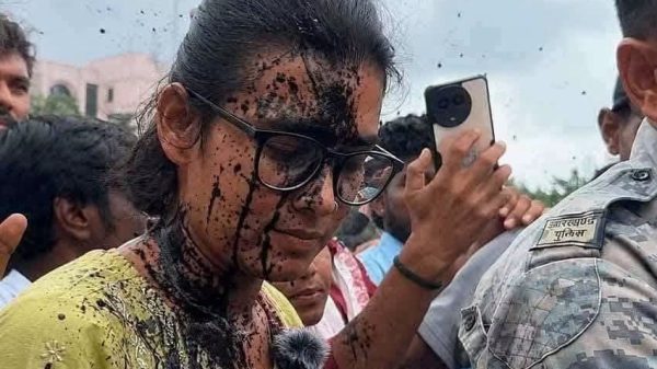 Young woman with glasses and a yellow shirt, mud splattered on her face and clothes, smiling in a crowd as someone takes a selfie nearby.