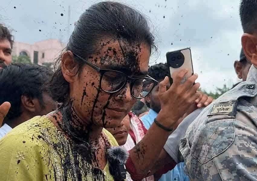 neha bora | No. 1 Indian Media News Portal Young woman with glasses and a yellow shirt, mud splattered on her face and clothes, smiling in a crowd as someone takes a selfie nearby.
