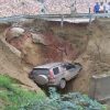 Car stranded in a collapsed hillside under a highway barrier, buried up to the wheels by mud and soil with bystanders nearby (informative image).